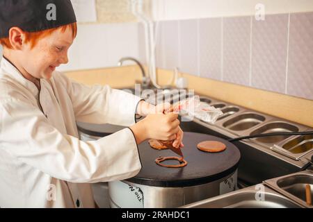 garçon cuisine des crêpes sur une crêperie professionnelle industrielle, cuisine dans le café, petit chef. Fait des cercles de pâte. rire en souriant. Banque D'Images