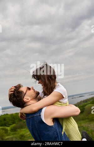 Vue latérale d'un homme souriant et branché en lunettes de soleil et gilet en denim qui embrasse et soulève une petite amie brune dans une sundress tout en passant du temps dans un décor rural Banque D'Images