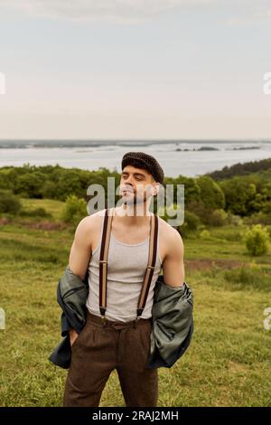 Homme tendance et barbu en veste, bretelles et casquette de jeune garçon tenant les mains dans les poches et regardant loin tout en étant de nature floue dans le dos Banque D'Images