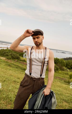 Homme barbu branché dans des bretelles touchant la casquette de jeune garçon et tenant la veste tout en regardant l'appareil photo avec paysage rural à l'arrière-plan, avant-mode moi Banque D'Images