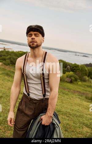 Portrait d'un homme à la mode et barbu dans des bretelles et une casquette de jeune garçon tenant une veste et regardant l'appareil photo tout en se tenant debout dans un paysage flou à la ba Banque D'Images