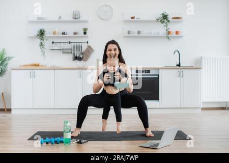 Adulte caucasien attrayant en vêtements de sport pratiquant des squats larges avec bébé dans les mains à l'intérieur de la cuisine. Jeune mère active engageant mignon bébé fille à des activités physiques à l'intérieur sur la journée ensoleillée. Banque D'Images