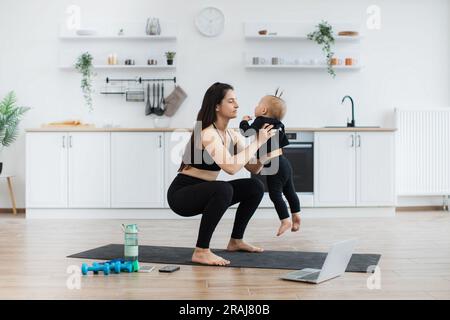 Adulte caucasien attrayant en vêtements de sport pratiquant des squats larges avec bébé dans les mains à l'intérieur de la cuisine. Jeune mère active engageant mignon bébé fille à des activités physiques à l'intérieur sur la journée ensoleillée. Banque D'Images