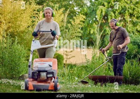 Jardinier masculin qui tond la pelouse avec un coupe-herbe tandis que son collègue utilise une tondeuse à gazon Banque D'Images