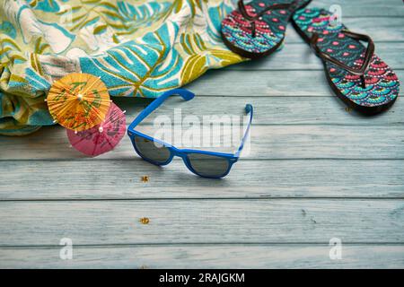 Lunettes de soleil bleues avec deux petits parapluies orange et rose, une serviette à imprimé tropical avec des tongs à imprimé tropical, sur le dessus d'une table en bois. Banque D'Images