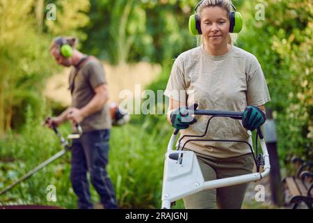 Jeune jardinière féminine utilisant une tondeuse à gazon pendant que son collègue ou ses patrons taillent le jardin en arrière-plan Banque D'Images