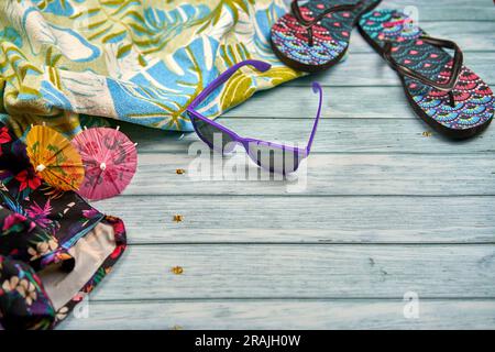 Lunettes de soleil violettes avec deux petits parapluies orange et rose, une serviette à imprimé tropical avec des tongs à imprimé tropical, sur une table en bois. Banque D'Images