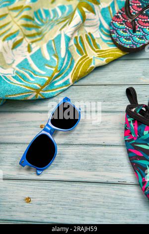 Lunettes de soleil bleues, tongs, maillot de bain et serviette de plage à imprimé tropical sur une table en bois. Banque D'Images