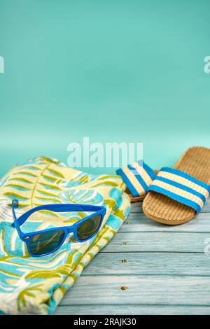 Nature morte estivale composée de lunettes de soleil bleues, sandales et serviette de plage, sur une table en bois et un fond vert. Banque D'Images