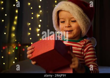 Un garçon de Noël en chapeau de père Noël souriant à la maison le soir. Nouvel an et jours fériés. Noël bonheur. Le garçon avec boîte cadeau s'amuser, profiter de Noël. Banque D'Images