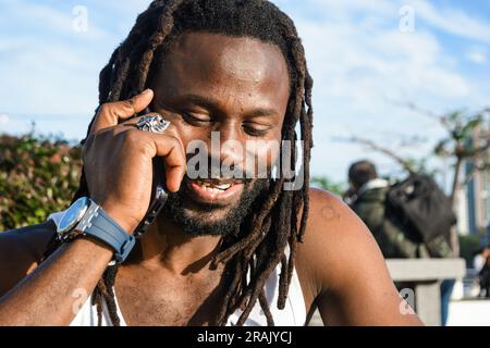 gros plan d'un jeune homme noir d'origine africaine, avec des dreadlocks et barbe, les yeux fermés souriant parlant au téléphone à l'extérieur, espace de copie. Banque D'Images