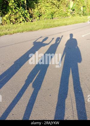 De longues silhouettes de trois amis jetées sur une route asphaltée pendant l’heure dorée, symbolisant l’unité et la joie dans un cadre extérieur. Banque D'Images