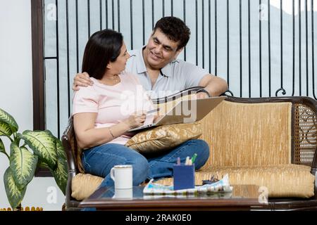 Portrait de jeune Indien heureux couple de famille joyeuse reposant sur le canapé dans le salon la recherche Internet tapant en ligne sur un ordinateur portable et parlant, Banque D'Images