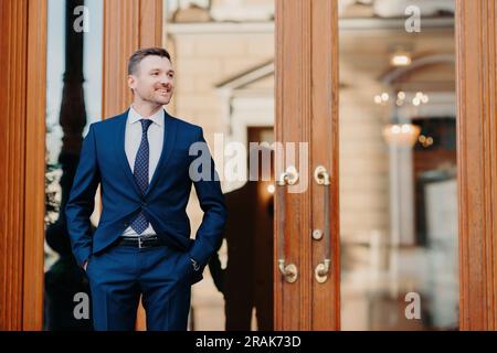 Travailleur satisfait en costume de luxe attend à l'entrée du restaurant, célèbre avec ses collègues, regarde joyeusement à part. Banque D'Images