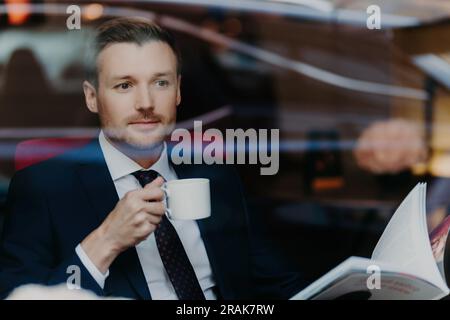 Beau patron dans un restaurant de luxe, tient la tasse, regarde à travers le menu, costume formel, profondément dans la pensée Banque D'Images