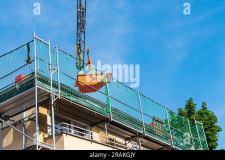 Grue de levage tient un conteneur orange au-dessus du toit d'une maison rénovée. La maille verte d'échafaudage enferment le toit. Banque D'Images