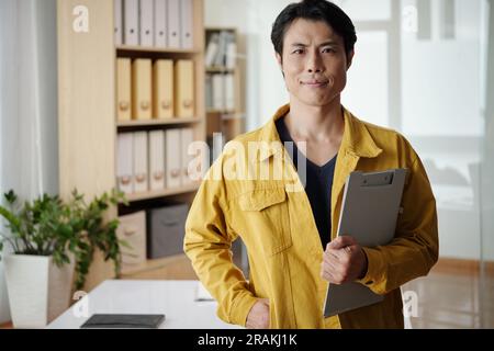 Portrait d'entrepreneur positif avec presse-papiers debout dans le bureau et souriant à la caméra Banque D'Images