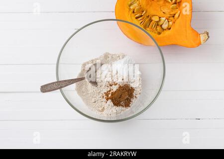 Bol en verre avec farine d'avoine, poudre à pâte et cannelle moulue, une demi-citrouille avec des graines sur un fond en bois blanc, vue de dessus. Stade de cuisson d Banque D'Images