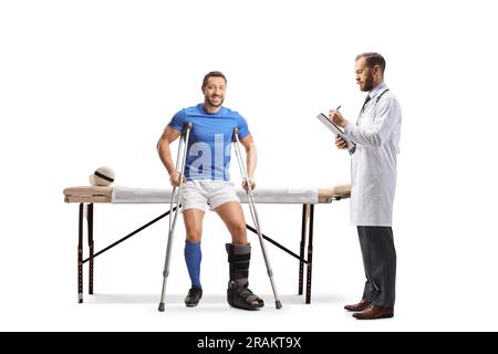 Joueur de football avec béquilles assis sur un lit de physiothérapie et un médecin écrivant un document isolé sur fond blanc Banque D'Images