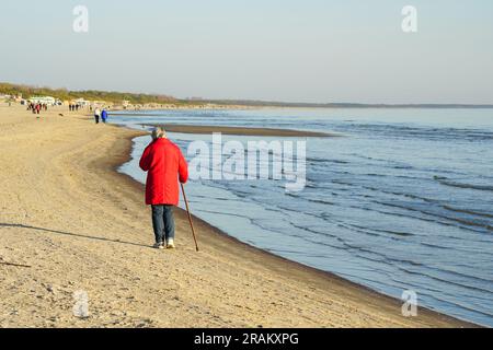 Une femme âgée dans une veste rouge avec un bâton de marche marche marche le long de la mer Baltique de sable Banque D'Images