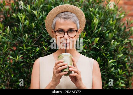 Femme mature élégante avec les cheveux gris buvant du matcha latte à l'extérieur Banque D'Images