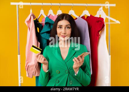 Femme shopaholic tenant le téléphone et la carte de crédit posant près du porte-vêtements, étant triste après des achats mobiles infructueux Banque D'Images