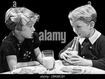 États-Unis : c. 1940. Un jeune garçon et une fille se regardant les uns les autres pendant qu'ils boivent du lait à la table du déjeuner. Banque D'Images