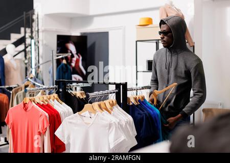 Homme afro-américain voleur prenant un risque, regardant des vêtements à la mode avant de le voler dans une boutique moderne. Le voleur portant des lunettes de soleil et une capuche ne doit pas être reconnu dans le centre commercial. Concept de cambriolage Banque D'Images