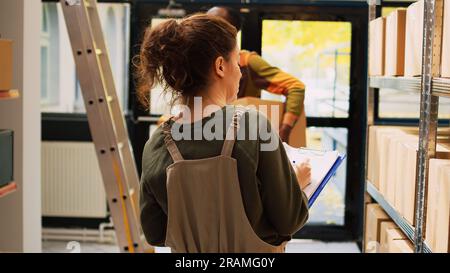Responsable du dépôt vérifiant la liste des produits dans la salle de stockage, travaillant sur la logistique et l'inventaire des stocks avec des boîtes sur des racks. Employé féminin analysant la marchandise de cargaison pour le contrôle de la qualité. Banque D'Images