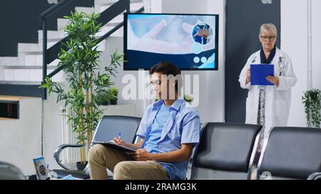 Patient assis sur chiar dans le hall de l'hôpital, remplissant les documents médicaux avec l'infirmière avant l'examen d'adaptation. Jeune adulte asiatique signant le formulaire d'assurance lors de la visite de contrôle. Service de médecine Banque D'Images