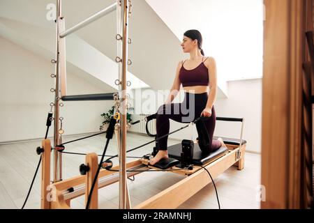 Une jeune fille fait Pilates sur un lit reformeur dans un studio lumineux. Un brunette mince dans un bodysuit bordeaux fait des exercices pour renforcer les bras an Banque D'Images