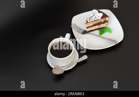 Une tasse de café noir sur une soucoupe avec une cuillère et une assiette avec un morceau de gâteau en couches de chocolat avec une fourchette et de la menthe sur un fond noir. Gros plan Banque D'Images