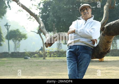 Vieil homme gai indien en bonne santé dans le parc debout à côté de l'arbre ayant du bon temps. Il porte une casquette de golf debout à côté d'une branche d'arbre. Banque D'Images