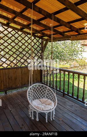 Une chaise hamac blanc est accrochée sur la terrasse en bois d'une maison privée. Banque D'Images