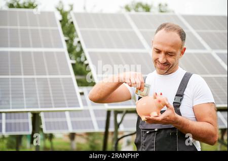 Homme adulte faisant des investissements dans l'avenir. Homme compétent investissant dans des sources d'énergie alternatives. Jeune homme mettant de l'argent dans une tirelire sur fond de panneaux solaires. Banque D'Images