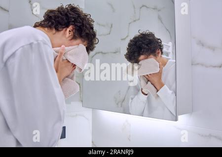 Jeune homme dans la salle de bains près du miroir lave le visage, essuyant avec une serviette Banque D'Images