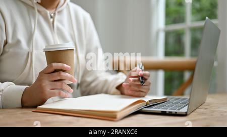 Image rapprochée d'une jeune femme asiatique en pull confortable sirotant un café et faisant ses devoirs dans un café. Banque D'Images