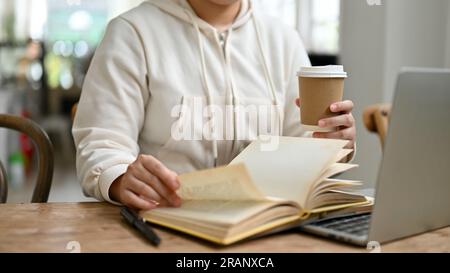 Image recadrée d'une femme en vêtements décontractés recherchant des informations dans un livre et sirotant un café tout en travaillant à distance dans un café le week-end. Banque D'Images