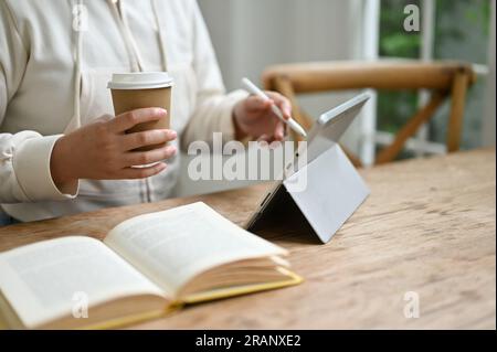 Image rapprochée d'une jeune étudiante asiatique dans des vêtements décontractés sirotant un café et travaillant sur son projet scolaire sur sa tablette numérique dans un c Banque D'Images