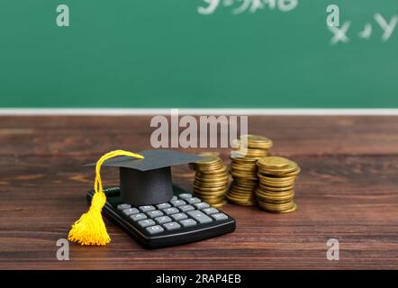 Casquette de graduation avec calculatrice et piles de pièces de monnaie sur table en bois brun contre tableau noir. Concept de prêt étudiant Banque D'Images