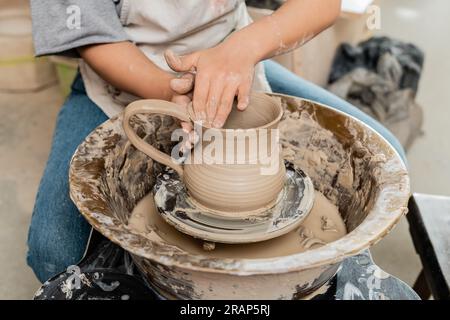 Vue recadrée de jeune artiste féminine en tablier créant cruche en argile sur la roue de poterie sur la table tout en travaillant dans l'atelier de céramique, poterie artisanale producti Banque D'Images