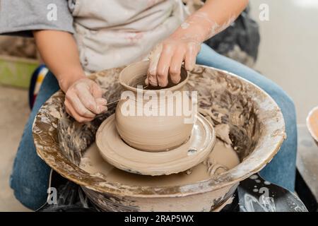 Vue recadrée de brune femme artisan en tablier coupant vase en argile sur la roue de poterie tout en travaillant dans l'atelier de céramique à l'arrière-plan, la mise en forme de l'argile et Banque D'Images