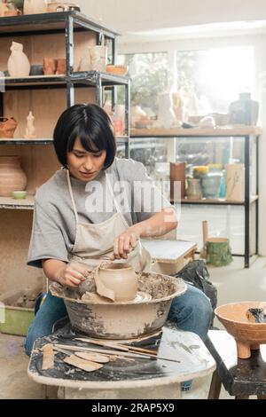 Brunette asiatique femme artisan en tablier coupant l'argile humide sur la roue de poterie en rotation tout en travaillant près des outils en bois et bol dans l'atelier de céramique floue Banque D'Images