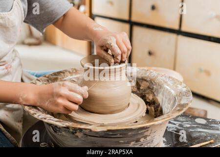 Vue recadrée de la jeune femme artisan en tablier coupant le vase d'argile humide sur la roue de poterie en rotation sur la table dans l'atelier de céramique floue, la mise en forme de l'argile et f Banque D'Images