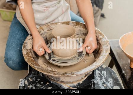 Vue recadrée de la jeune femme artisan en tablier coupant vase d'argile sur la roue de poterie en rotation près du bol avec de l'eau à l'arrière-plan dans l'atelier de céramique, arti Banque D'Images