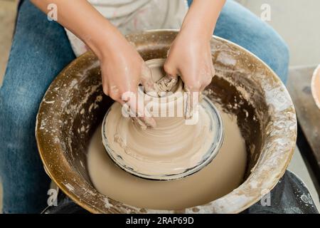 Vue à angle élevé de jeune potier femelle en tablier façonnant l'argile humide tout en travaillant sur la roue de poterie en rotation dans l'atelier d'art à l'arrière-plan, potier qualifié Banque D'Images