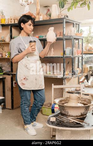 Jeune artiste féminine asiatique en tablier tenant sculpture et café pour aller près de l'argile sur la roue de poterie, des outils et rack dans l'atelier de céramique, la création artisanale Banque D'Images