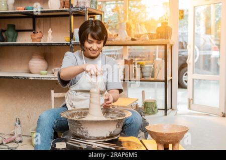 Souriant jeune artiste féminine asiatique en tablier versant de l'eau sur l'argile sur la roue de poterie près des outils et bol dans l'atelier flou au coucher du soleil, artisan création Banque D'Images