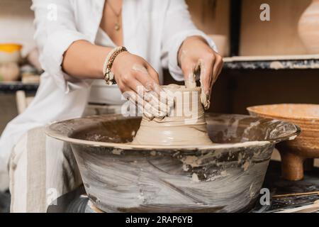 Vue recadrée de la jeune femme artisan en vêtements de travail façonnant l'argile humide tout en travaillant sur la roue de poterie près du bol à l'arrière-plan, espace de travail de studio de poterie et Banque D'Images