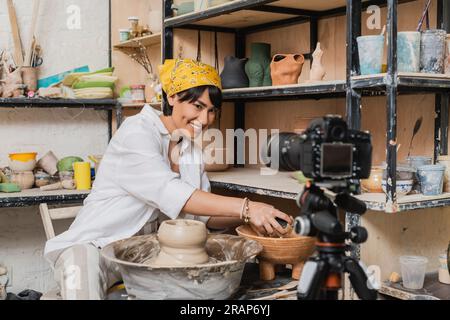 Artisan asiatique souriant en foulard tenant l'éponge près du bol avec l'eau, l'argile et la roue de poterie et regardant l'appareil photo numérique dans l'atelier d'art, argile Banque D'Images
